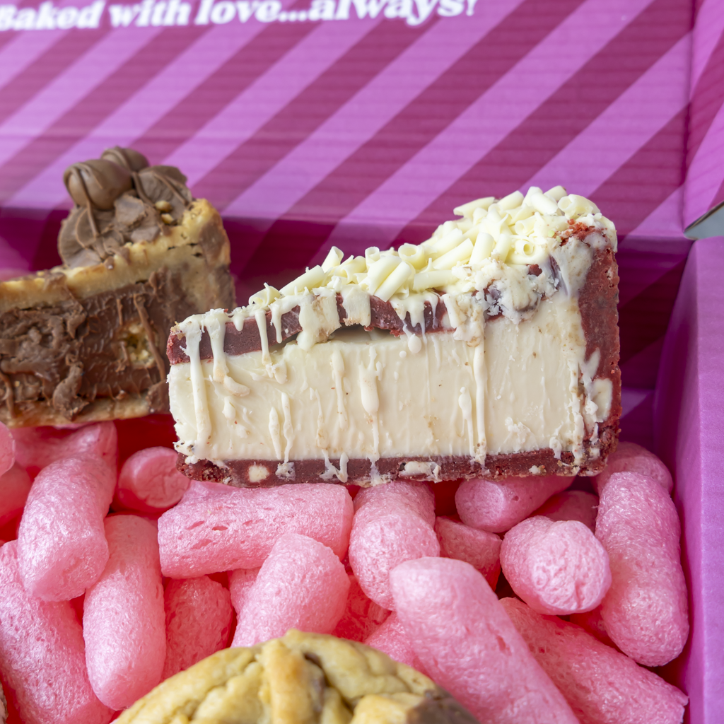 A close-up of a slice of chocolate and cream red velvet cookie pie slice with white chocolate drizzle, resting on pink packing gift box with other baked goods partially visible.
