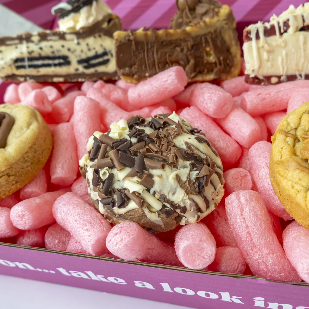 A close-up of assorted chocolate-covered cookies and bars arranged in pink packing peanuts inside a purple box. The center cookie is topped with white chocolate and chocolate shavings, with other decorated sweets visible around it.