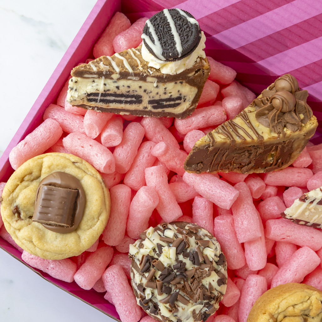 A close-up of assorted novelty chocolate cookie pie slices and cookies arranged in pink packing inside a pink striped DoughGirl box.