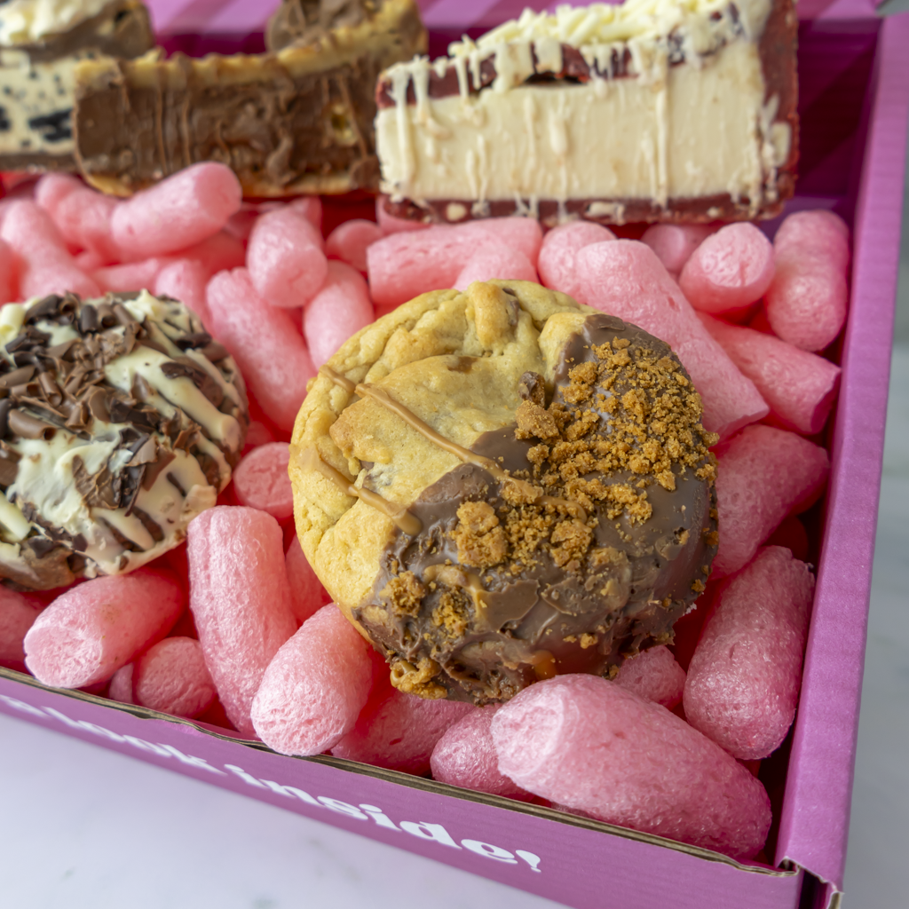 A close-up of assorted chocolate-covered cookies or pastries arranged inside a pink box filled with pink packing peanuts. The center pastry is golden with dark chocolate and cookie crumb topping, while other pastries are partially visible in the background.