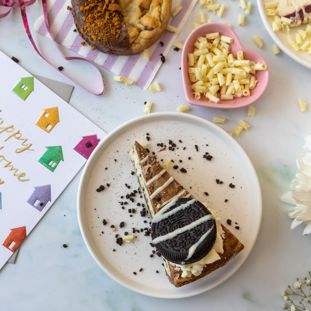 A slice of chocolate cake with white chocolate drizzle and an Oreo cookie on a white plate, styled on a marble surface with a heart-shaped dish of white chocolate curls, a cookie, and a 'Happy Home' card.