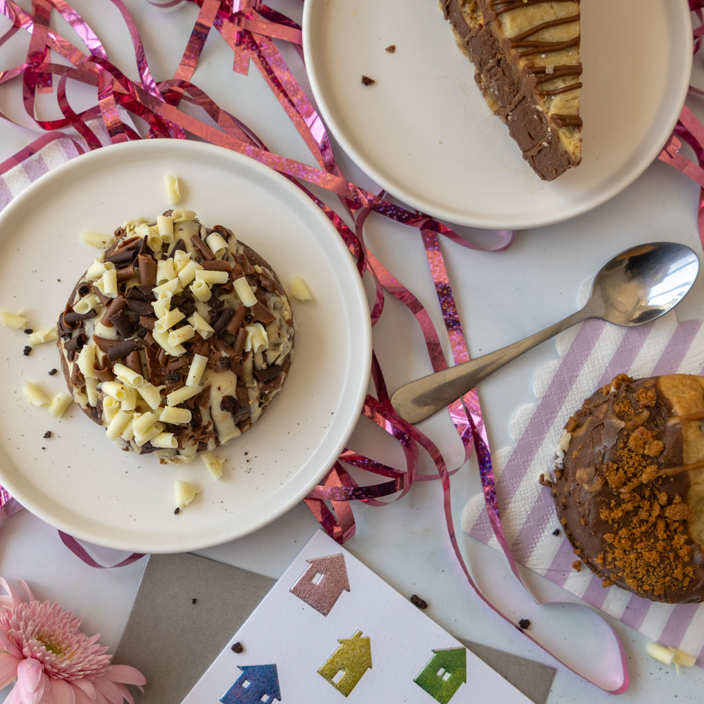 A styled flat-lay of chocolate desserts on white plates, including a round chocolate cookie topped with white chocolate shavings and a slice of chocolate cookie pie with caramel drizzle, alongside a spoon, pink ribbon streamers, and a small flower on a white surface.