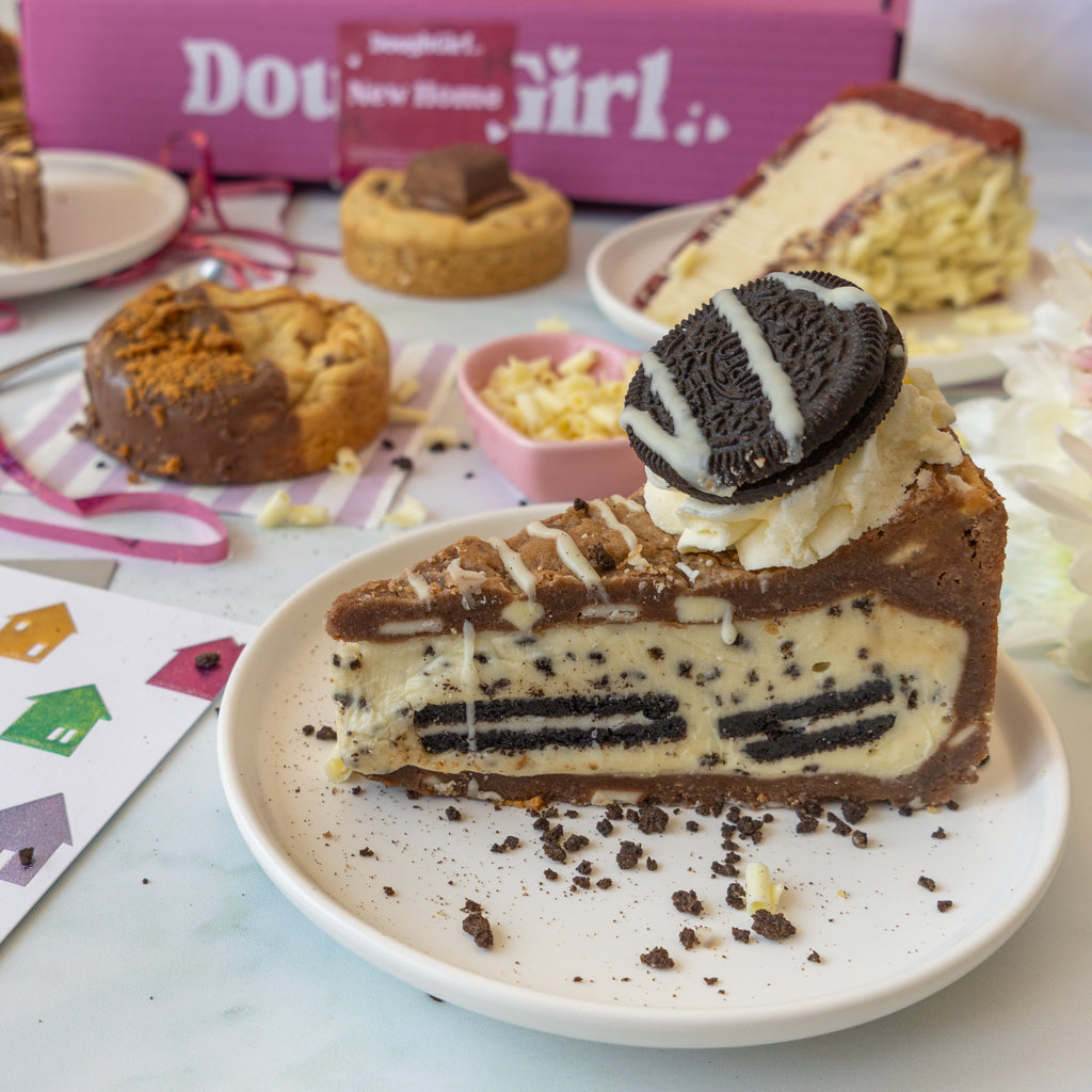 A slice of Oreo-themed cookie pie slice on a white plate, topped with a whole Oreo cookie, with other cake slices and a pink branded box in the background.