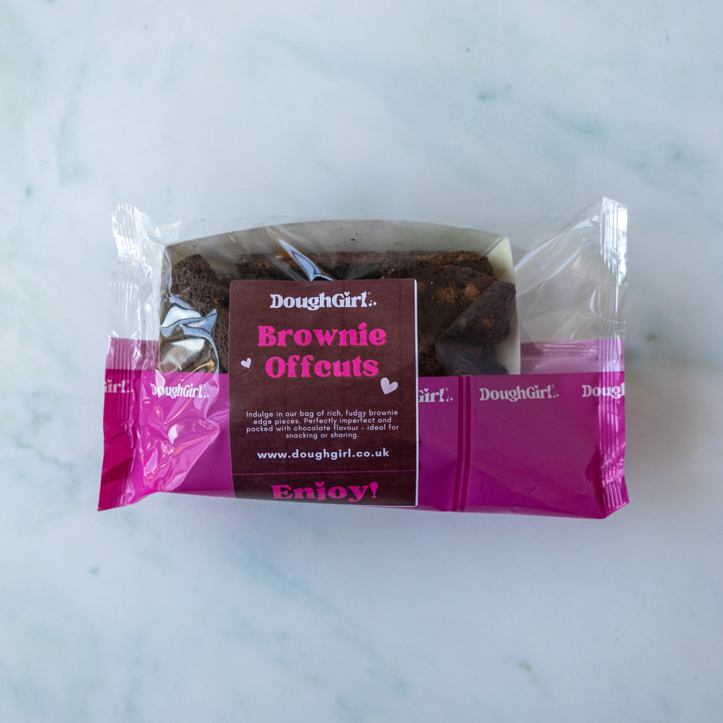 A packaged brownie offcut from Doughgirl, sealed in clear plastic with a bright pink and dark brown paper label, placed on a light marble surface.