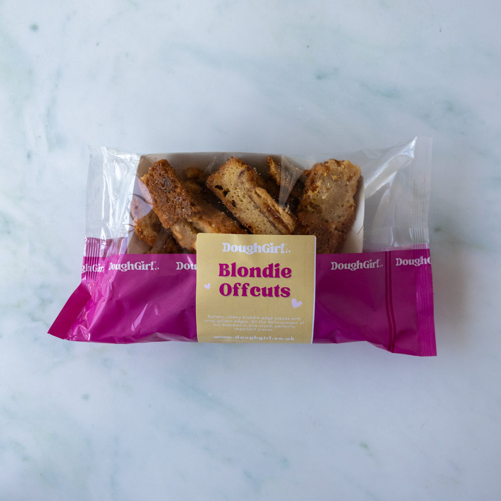 A package of Doughgirl Blondie Offcuts containing several golden-brown baked biscuit pieces, sealed in a clear plastic bag with a bright magenta and cream paper label, set on a light marble surface.
