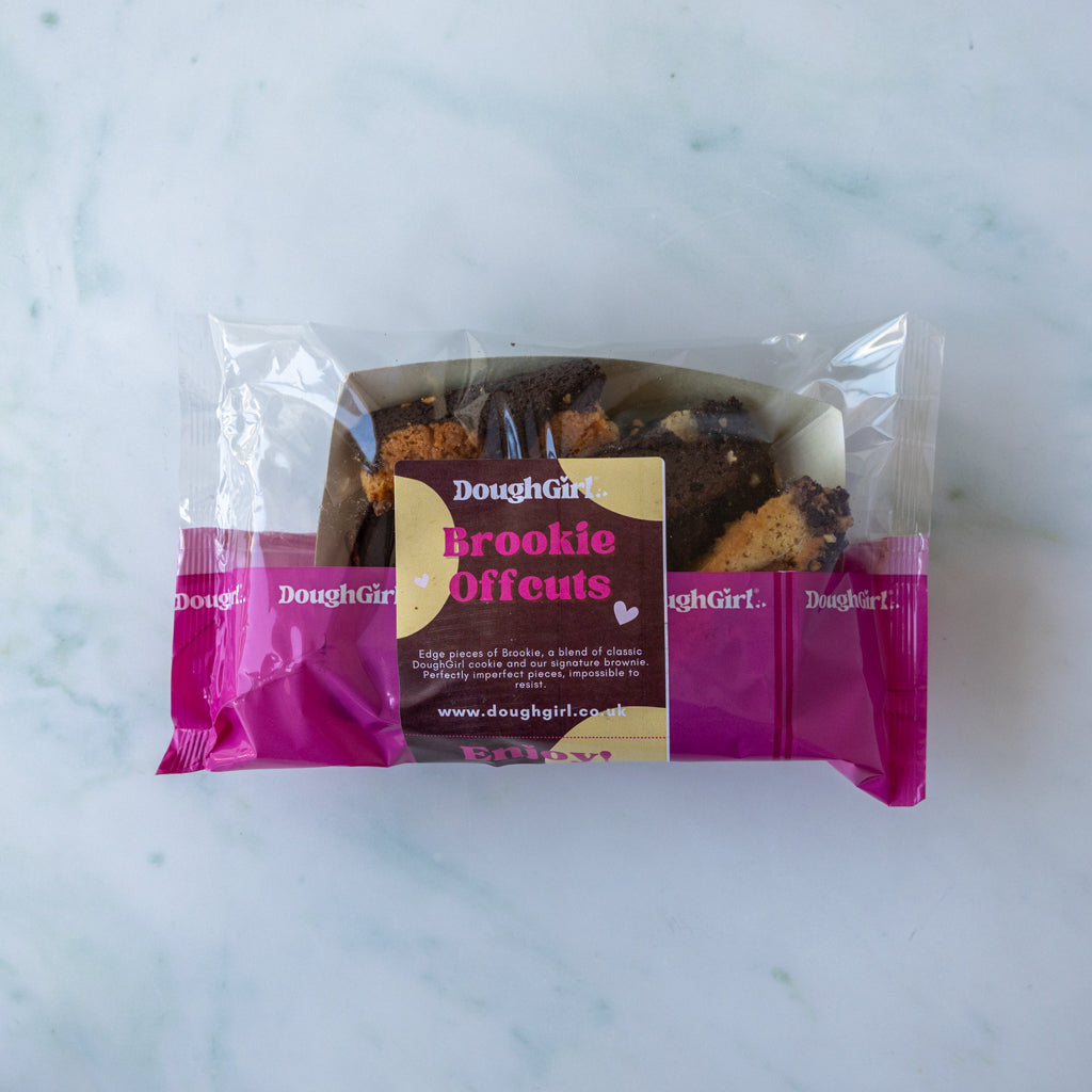 A package of DoughGirl Brookie Offcuts cookies on a light marble surface. The product is in a clear plastic bag with a bright magenta wrapper featuring a central cream and brown label.