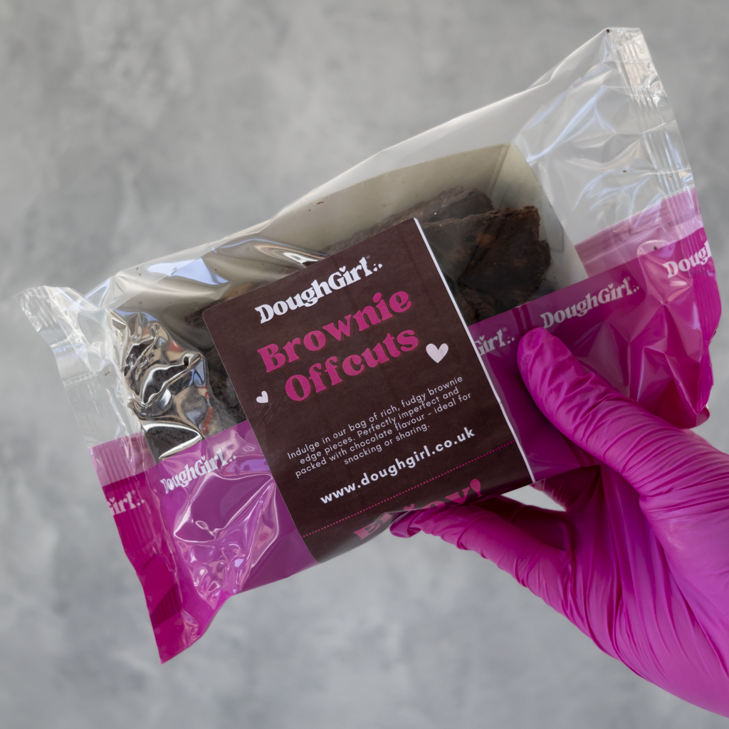 A hand wearing a bright pink glove holds a clear plastic bag containing dark brown baked brownies with a branded brown paper label that reads DoughGirl Brownie Offcuts.