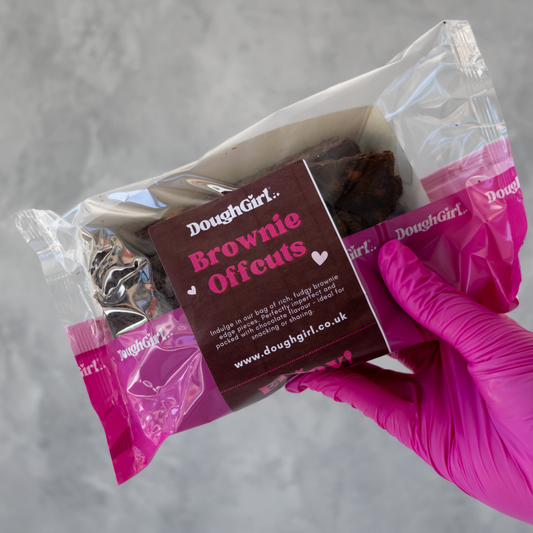 A hand wearing a bright pink glove holds a clear plastic bag containing dark brown baked brownies with a branded brown paper label that reads DoughGirl Brownie Offcuts.