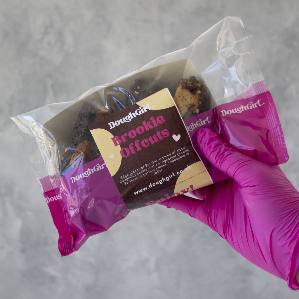 A hand wearing a bright pink glove holds a clear plastic bag containing 'Brookie Offcuts' from Doughgirl. The packaging features a prominent brown and beige label with the Doughgirl brand name.
