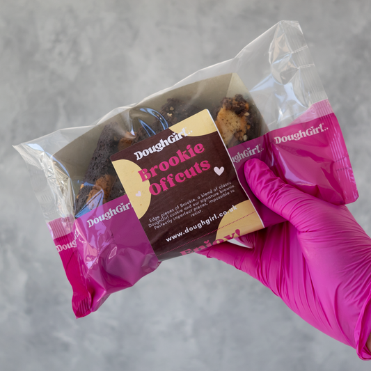 A hand wearing a bright pink glove holds a clear plastic bag containing 'Brookie Offcuts' from Doughgirl. The packaging features a prominent brown and beige label with the Doughgirl brand name.
