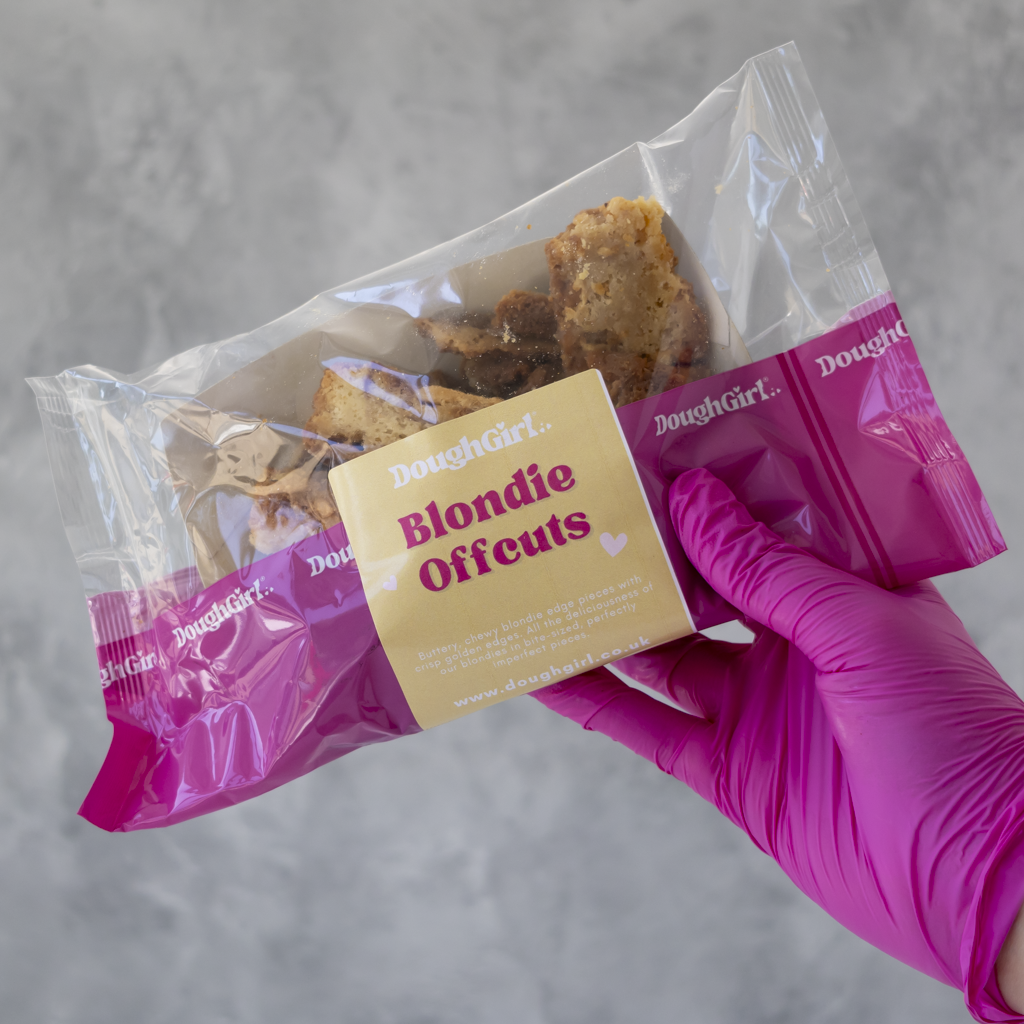 A hand wearing a bright pink glove holds a clear plastic bag of DoughGirl Blondie Offcuts