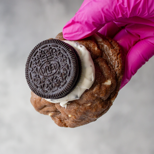 Hand wearing a pink glove holding a Oreo Deluxe Cookie with an Oreo sandwiched inside against a neutral background