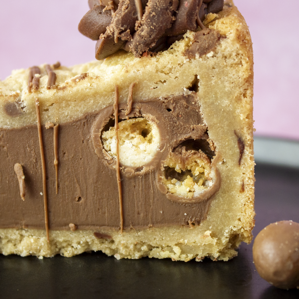 Close-up of a slice of chocolate chip cookie cake with a Malteser chocolate ball on a pink background