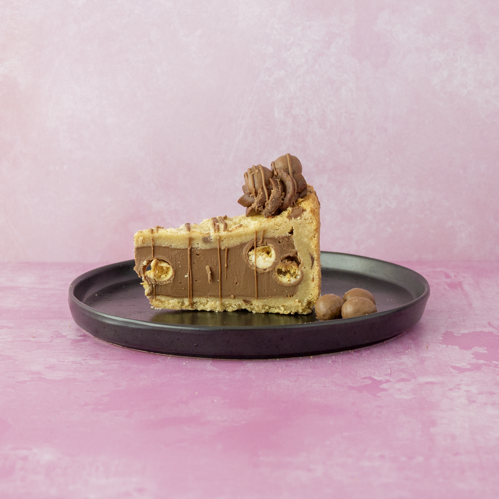 Slice of Full Malteser Cookie Pie with malteser chocolate balls on a black plate against a pink background