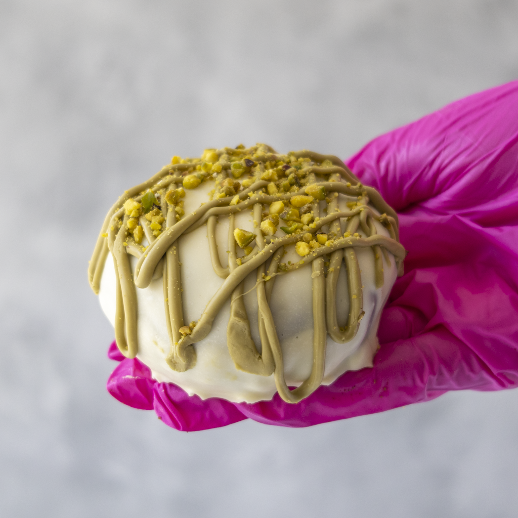 DoughGirl Holy Pistachioli Choccy Boom Bite held in pink glove with creamy white chocolate coating