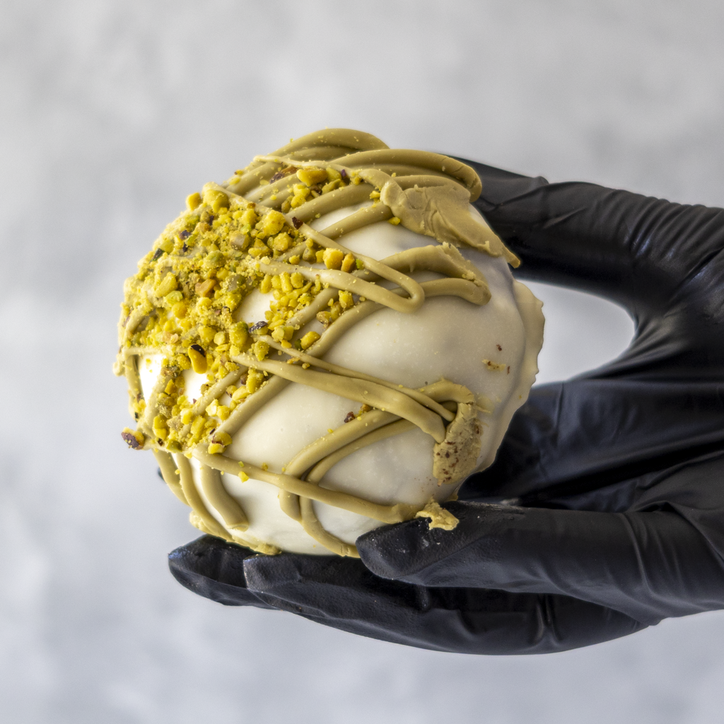 Holy Pistachioli Choccy Boom Bite held in black glove showing pistachio drizzle and nut topping