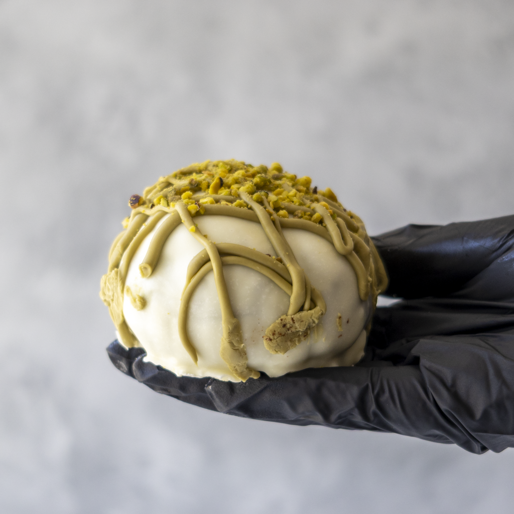Holy Pistachioli Choccy Boom Bite held in black glove with white chocolate coating and pistachio topping