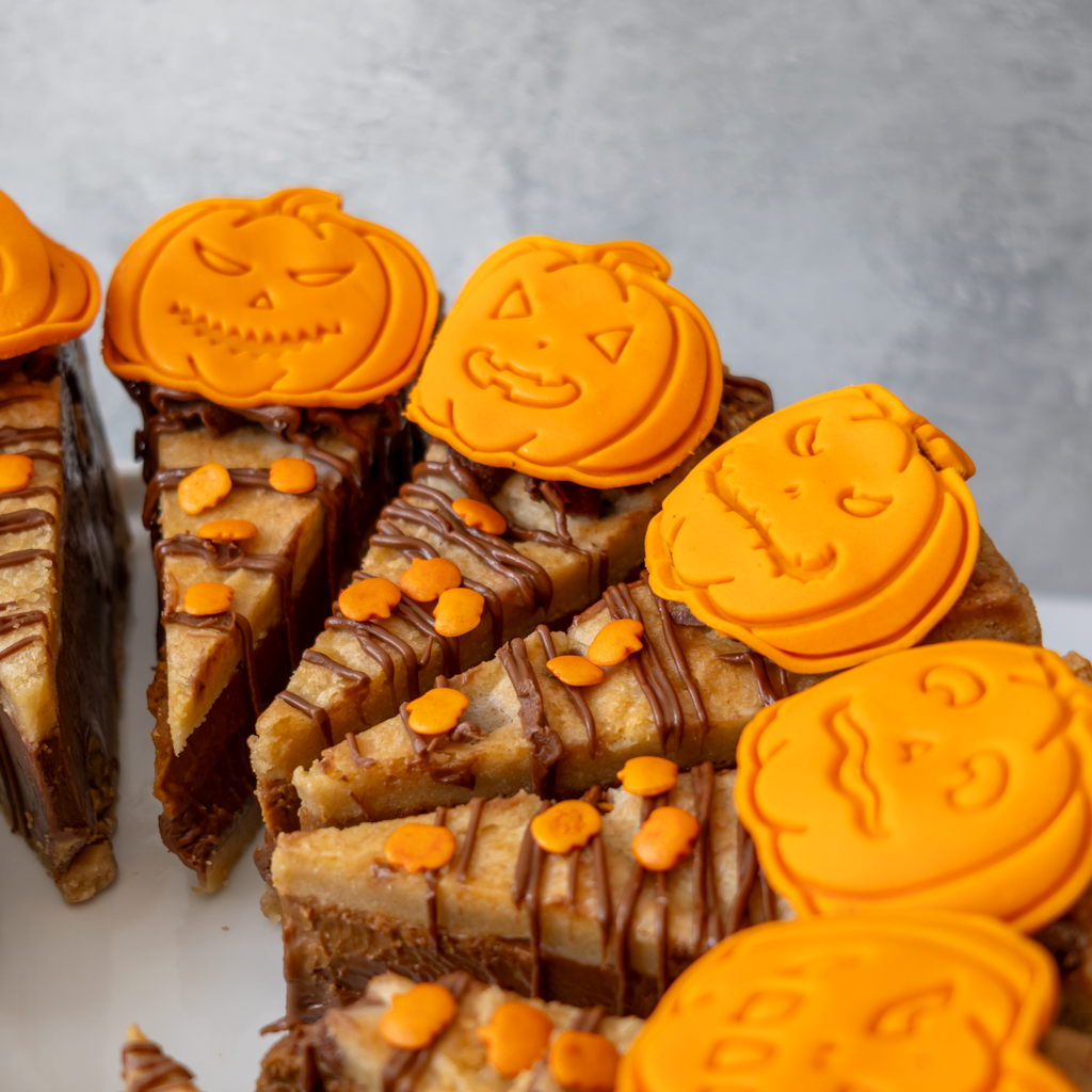 Halloween-themed pumpkin cookie pie arranged on a white plate, featuring orange pumpkin-shaped toppings and small orange candy sprinkles, with some bars sliced to show a layered dessert filling.