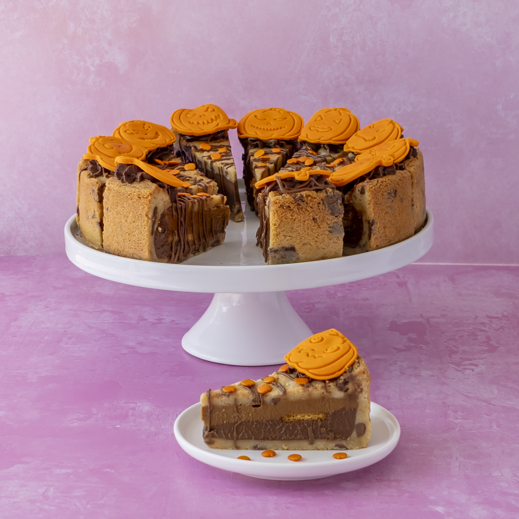 A layered full Pumpkin Patch Cookie Pie with orange caramel swirls, presented on a white cake stand with a slice placed below on a small plate, set against a pink background.