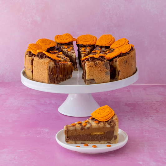 A layered full Pumpkin Patch Cookie Pie with orange caramel swirls, presented on a white cake stand with a slice placed below on a small plate, set against a pink background.