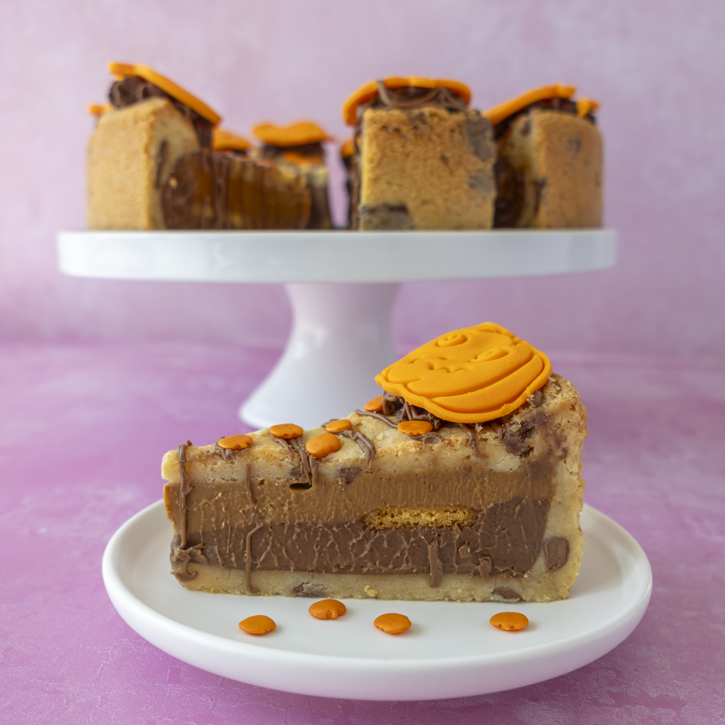 A slice of layered pumpkin spice and chocolate cookie pie with orange drizzle and candy decorations sits on a white plate in the foreground, with a white cake stand holding several slices in the background against a pink backdrop.