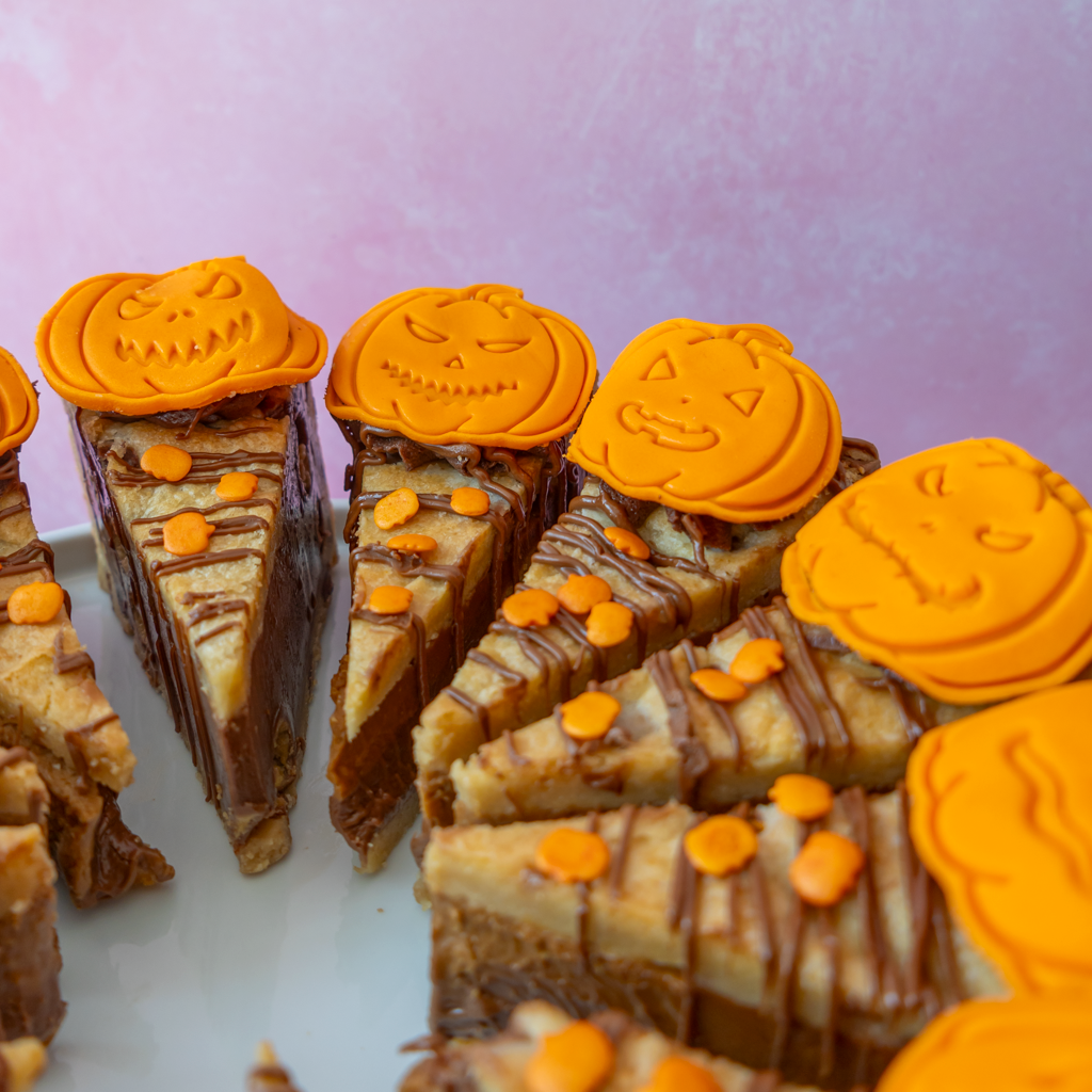 Halloween-themed pumpkin cookie pie slices decorated with orange pumpkin-shaped toppings and chocolate drizzle, arranged on a white plate against a soft pink background.