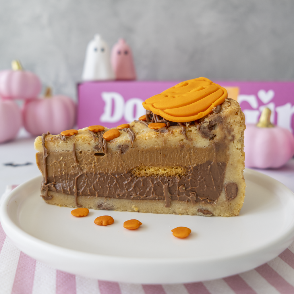 A slice of Pumpkin Patch Cookie Pie on a white plate, decorated with a pumpkin-shaped topping and orange sweets and chocolate drizzle, with a pink DoughGirl box and small pumpkins in the background.