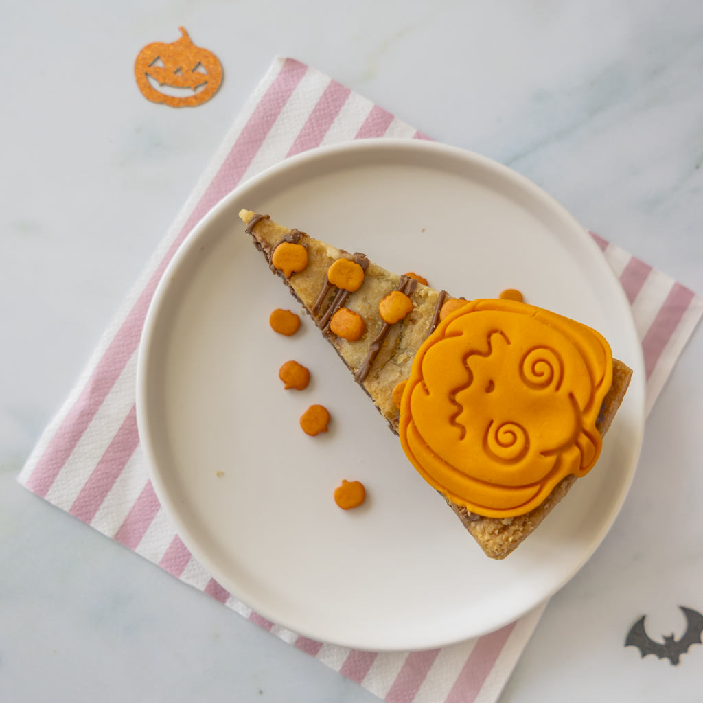 A Halloween-themed slice of cookie pie on a white plate, decorated with orange pumpkin-shaped frosting and small orange sweet pieces, set on a pink and white striped napkin with pumpkin and bat decorations.
