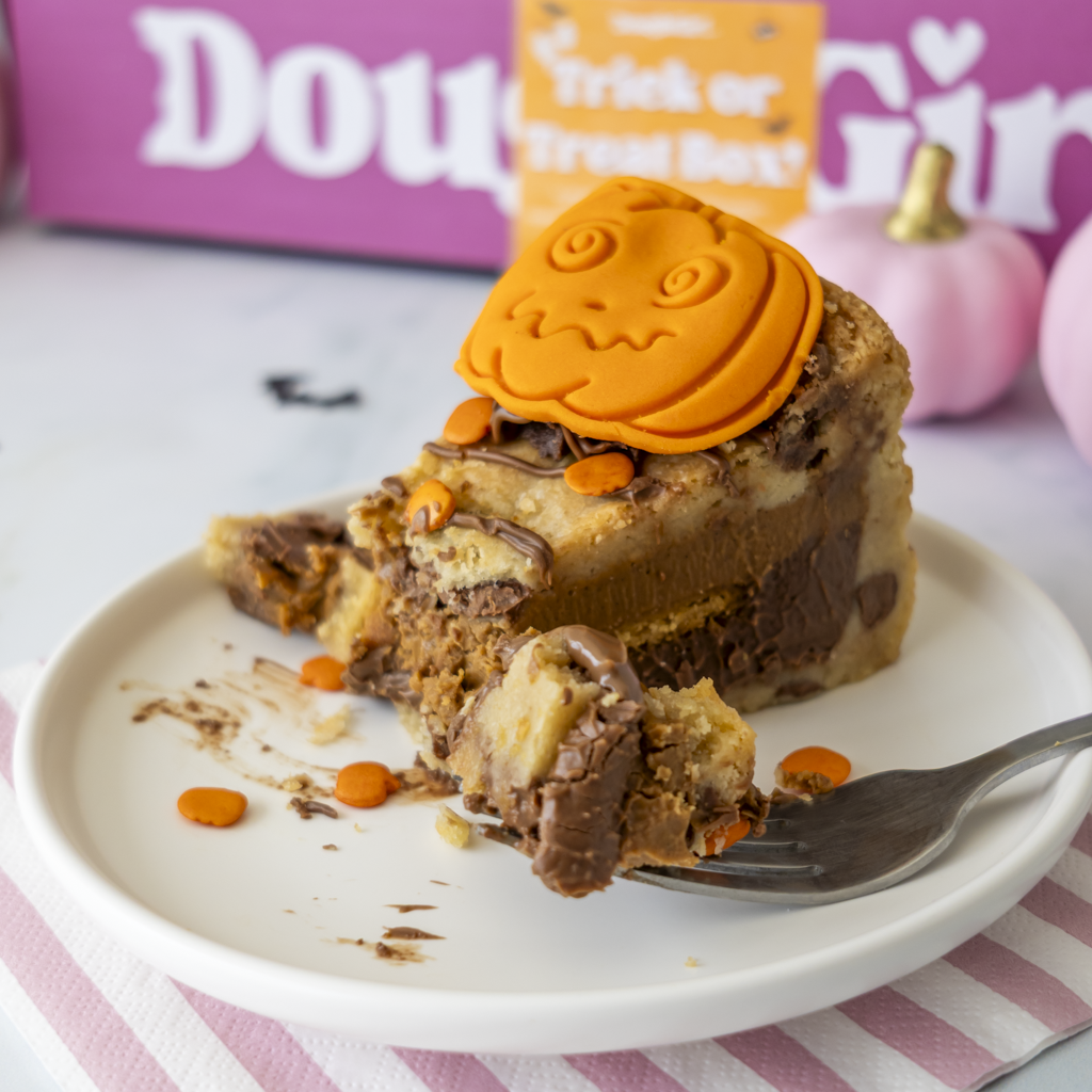 A close-up of a slice of layered chocolate and cookie pie on a white plate with a fork, topped with an orange pumpkin decoration. The background shows a pink 'DoughGirl' box and a 'Trick or Treat Box' sign with small pumpkins.