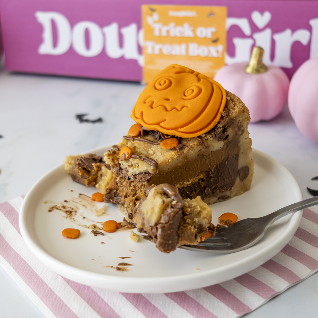 A slice of layered Pumpkin Patch Cookie Pie topped with an orange pumpkin-shaped fondant decoration and orange candy corns, served on a white plate with a fork. A pink and white 'DoughGirl' box and a small orange 'Trick or Treat Box' card are visible in the background with pink pumpkins.