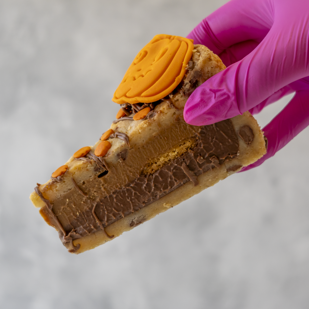 A hand wearing a bright pink glove holds a thick slice of layered Pumpkin Patch Cookie Pie Slice in a cookie crust with caramel drizzle, sweets, and a pumpkin-shaped topping against a soft grey background.