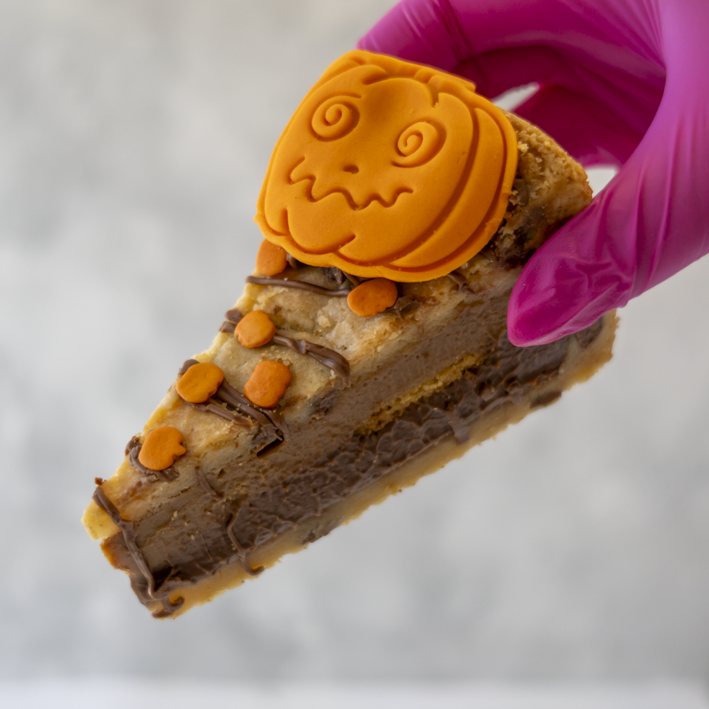 A close-up of a hand wearing a bright pink glove holding a slice of layered chocolate and cookie pie with orange pumpkin-shaped decorations on top, against a soft grey background.