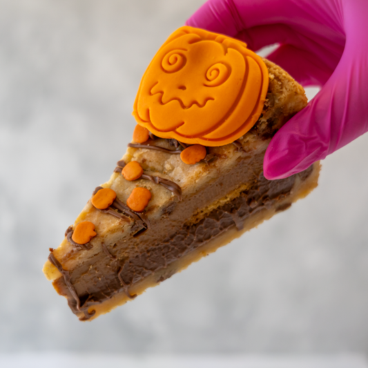 A close-up of a hand wearing a bright pink glove holding a slice of layered chocolate and cookie pie with orange pumpkin-shaped decorations on top, against a soft grey background.