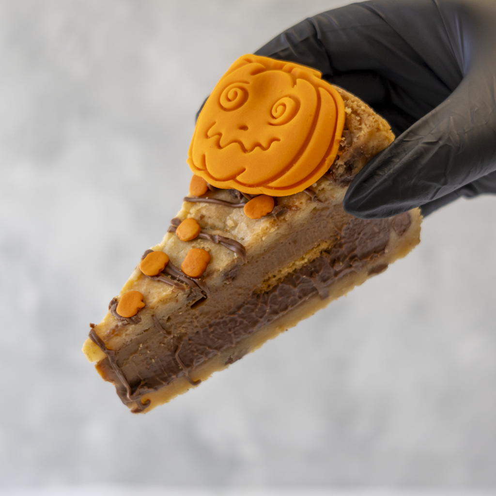 A close-up of a hand wearing a black glove holding a slice of Pumpkin Patch Cookie Pie decorated with orange pumpkin-shaped frosting and small orange candies, against a soft gray background.