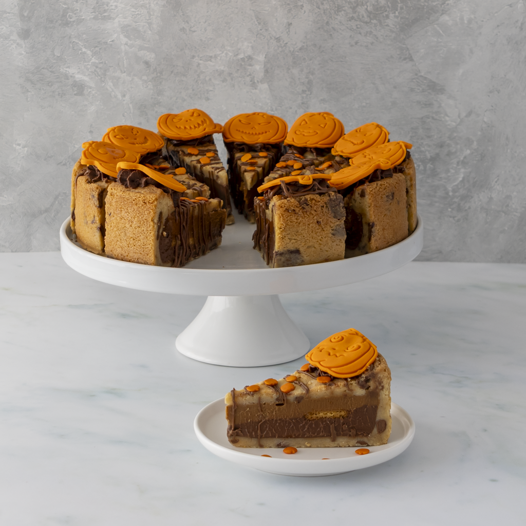 A layered chocolate and pumpkin spice cookie pie displayed on a white cake stand, with a single slice placed on a small plate in front. The cake is topped with orange circular decorations, set against a light grey textured wall and white marble surface.