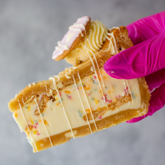 Slice of DoughGirl Party Popper Cookie Pie Slice with creamy white chocolate, colourful sprinkles and party rings, held by a hand wearing a pink glove against a gray background.