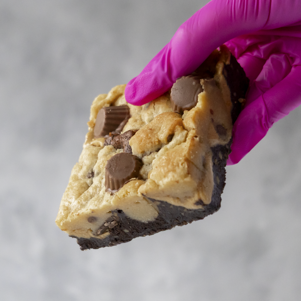 Peanut Overload Brookie Slab held by a hand wearing a pink glove on a gray background