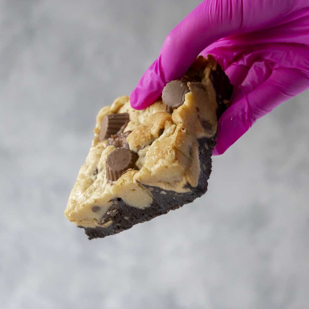 Hand wearing a pink glove holding a layered Peanut Overload Brookie Slab against a gray background
