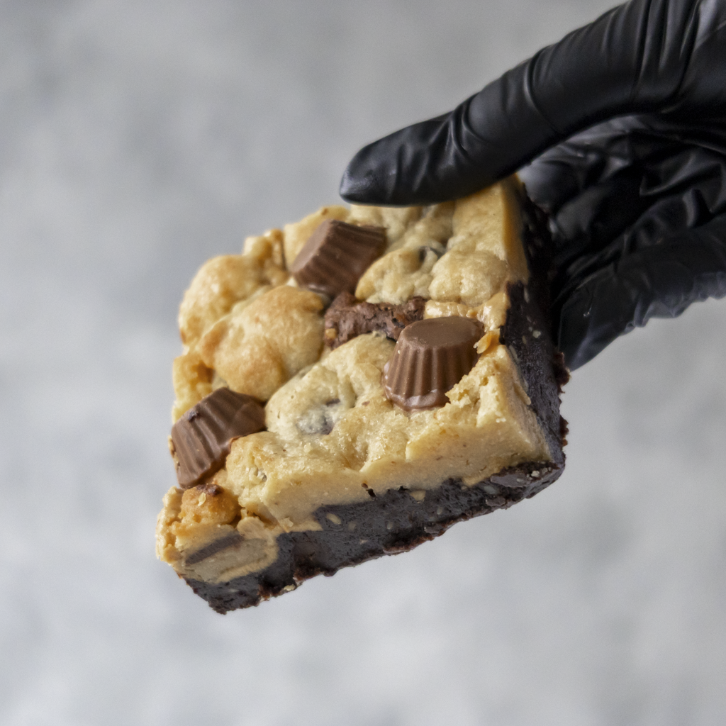 Hand holding a Peanut Overload Brookie Slab by DoughGirl UK with chocolate and peanut butter cups against a neutral background.