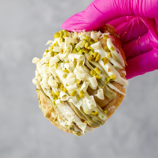 Hand holding a Pistachio & White Chocolate Stuffed Deluxe Cookie with white frosting and green pistachio pieces, against a gray background.