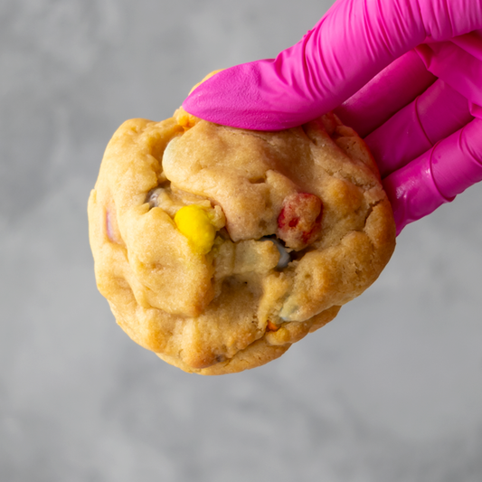 Rainbow Deluxe Cookie held by a hand wearing a pink glove against a gray background