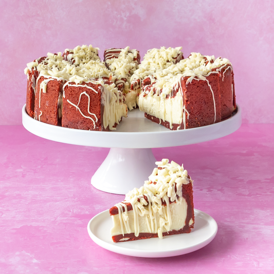 A Red Velvet Cookie Full Pie with white chocolate drizzle sits on a white pedestal stand, with one slice placed on a small plate in front. The background is a soft pink studio backdrop.
