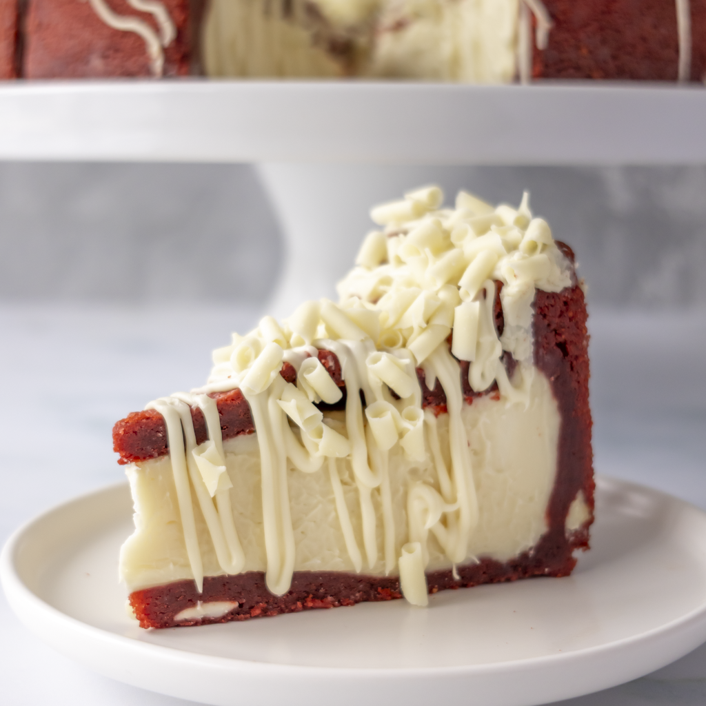 A slice of Red Velvet Cookie Pie with white chocolate drizzle on a white plate, with a cake stand and more cake blurred in the background.