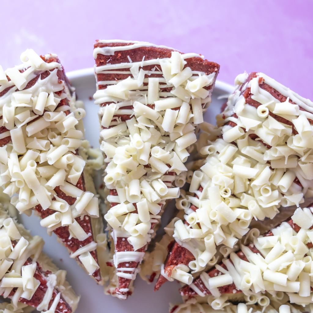 Close-up of several triangular slices of a red velvet cookie pie topped with white chocolate shavings and drizzles, arranged on a white plate against a pink background.