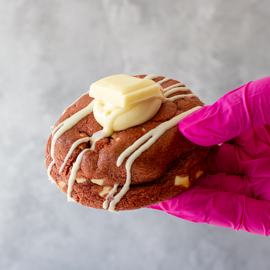 Red Velvet Deluxe Cookie with white icing held by a hand wearing a pink glove on a gray background
