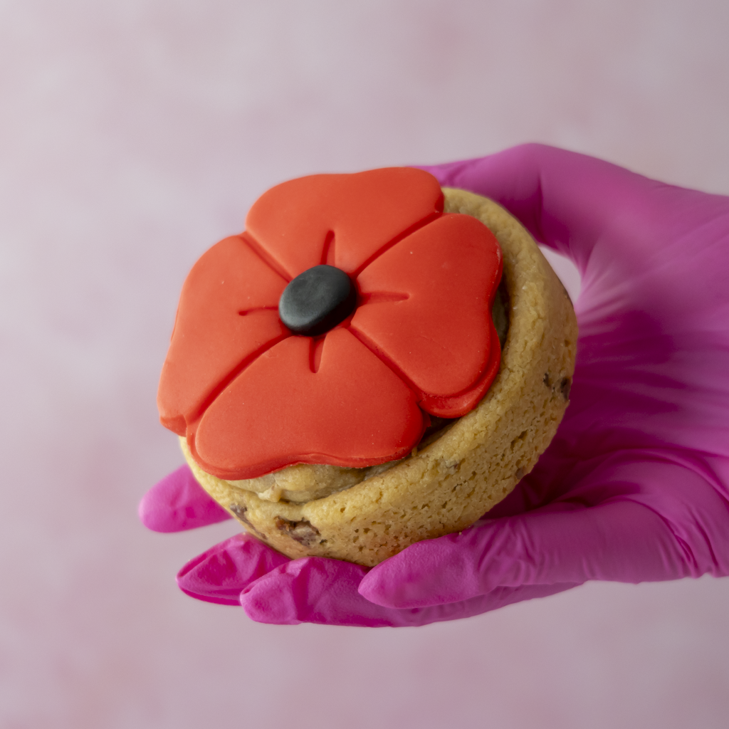 A hand wearing a bright pink glove holds a chocolate chip cookie topped with a large, vibrant red poppy flower decoration with a small black centre, set against a soft pink background.