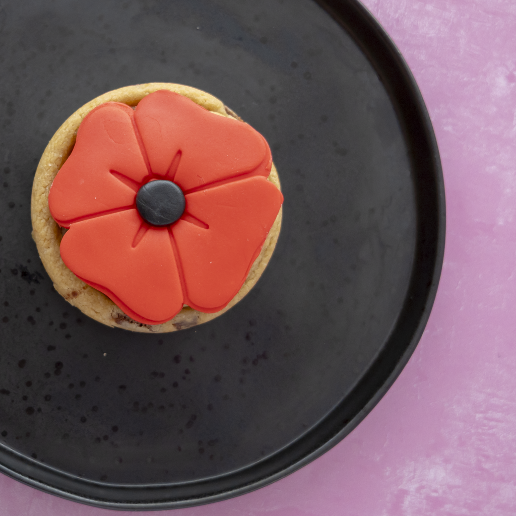 A small baked cookie topped with a bright red poppy flower decoration, placed on a black plate against a pink background.
