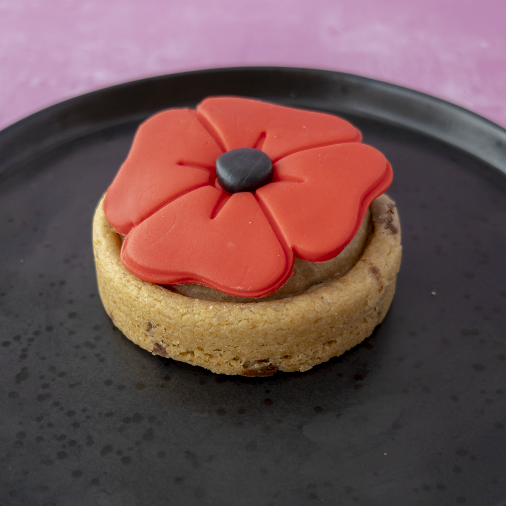 A round cookie topped with a bright red poppy flower decoration and a small black centre, presented on a dark plate against a pink background.