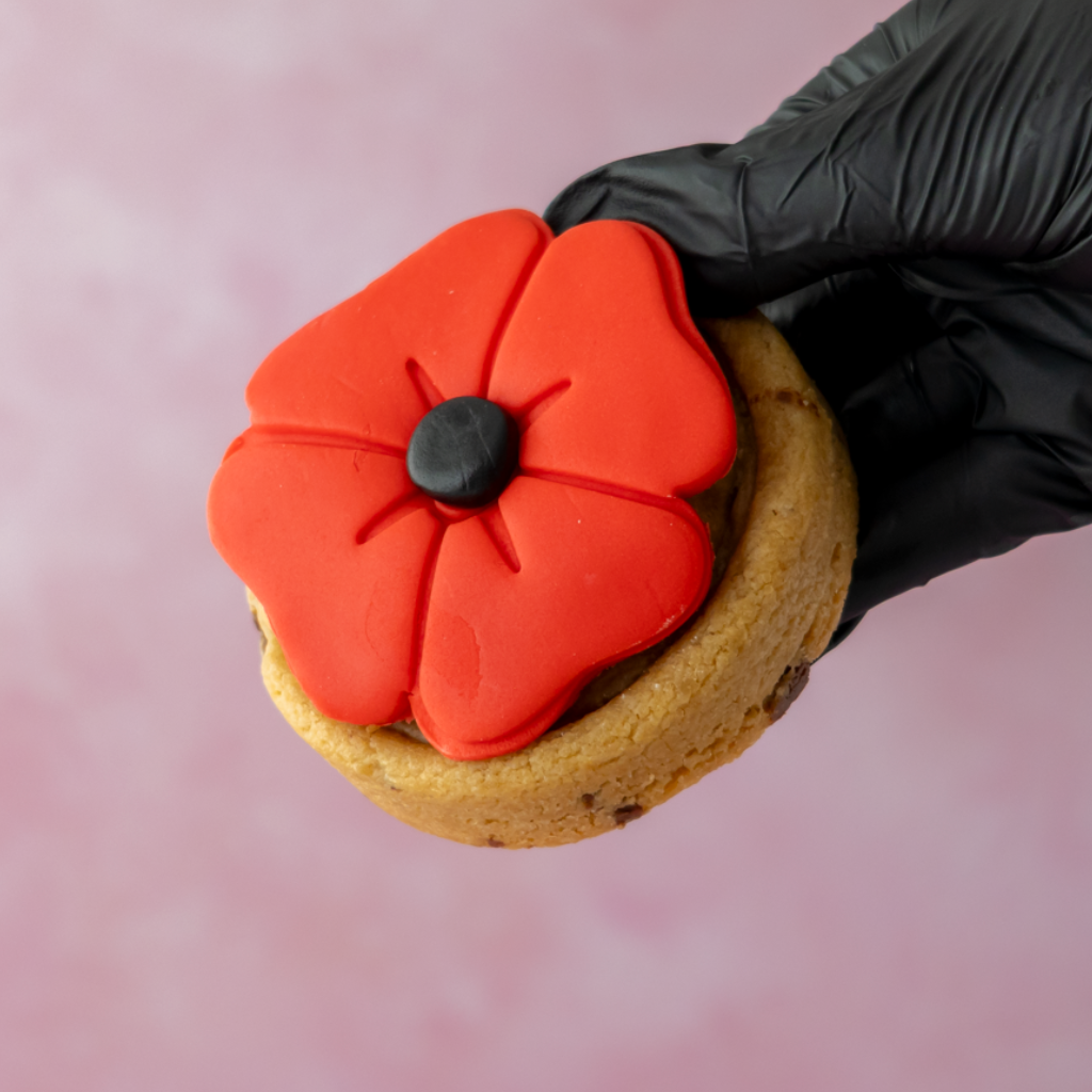 A hand wearing a black glove holds a decorated poppy-shaped cookie against a soft pink background. The cookie has a golden-brown base topped with bright red poppy petals and a small black center.
