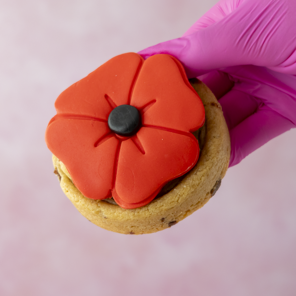 A close-up of a hand wearing a bright pink glove holding a DoughGirl cookie topped with a large red poppy flower decoration and a small black centre, against a soft pink background.